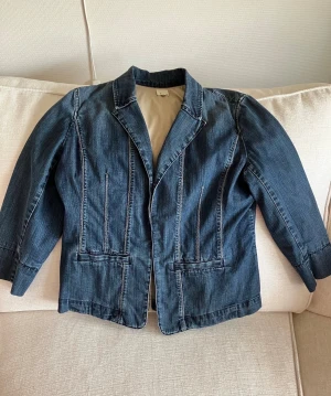 Blå jeanskavaj från Ecru - Snygg blå jeanskavaj från Ecru med klassisk krage och dekorativa sömmar. Jackan har två fickor fram och är tillverkad i jeansmaterial med en cool, figurnära passform. Perfekt för dig som gillar en avslappnad men stilren look 💙          Ärmarna och halva ryggen är fodrade. Mjuk men slittålig kvalitet. Längd bak : 58 cm Ärmlängd : 50 cm Knäpps med 2 öglor på insidan : bild 6