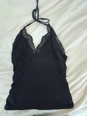 Svart spetsdetaljerad linnetopp - Svart linnetopp med spetsdetaljer och v-ringad hals. Toppen har tunna axelband som knyts i nacken och en croppad passform. Spetsen har blommönster och ger en snygg kontrast mot det släta tyget. Perfekt för dig som gillar stilrena och trendiga plagg.