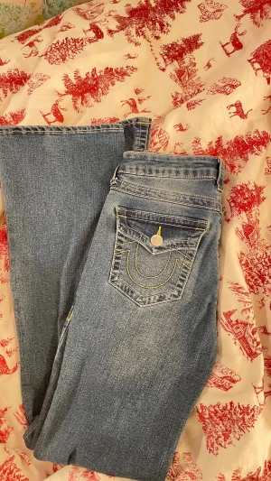 True Religion ljusblå jeans med kontrastsöm - Säljer ett par ljusblå jeans från True Religion med gul kontrastsöm och snygga detaljer på bakfickan.  De är aldrig använda. Modellen har raka ben och klassisk femficksdesign med silverfärgade knappar och logotyp på bakfickan. 