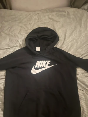 Svart Nike hoodie med vit logga - Svart hoodie från Nike i storlek M med stor vit Nike-logga på bröstet. Tröjan har huva och magficka, samt långa ärmar. Materialet är mjukt och känns som bomull och polyester, perfekt för chill dagar. Klassisk och enkel design som alltid funkar.