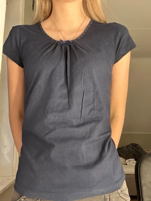 Mörkblå prickig blus från Isolde - Supersöt mörkblå blus från Isolde med vita prickar runt halsen och en liten rosett framtill. Blusen har korta ärmar och ett lätt rynkat parti vid halsen som ger en fin form. Perfekt för dig som gillar detaljer och klassisk stil.