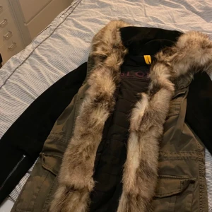 Parkas med fuskpäls från Blonde No.8 - Snygg parkas från Blonde No.8 i olivgrönt med svarta ärmar och stor huva med fluffig fuskpäls. Jackan har flera fickor, knappar framtill och quiltat innerfoder. Perfekt för kalla dagar och riktigt trendig look. Jackans dragkedja längst ner har släppt 3 cm från tyget men är åtgärdat hos skomakaren sommaren 2024 och sitter på sin plats och är kontrollerad innan försäljning! annars finns det inga defekter eller dylikt på jackan. 