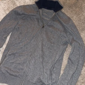 Grå kofta med dragkedja - Snygg grå kofta med hel dragkedja och ribbad mörkblå krage. Tröjan har långa ärmar och ribbade muddar vid ärmslut och nederkant. Perfekt för dig som gillar enkel och stilren design. Passar till jeans eller chinos för en chill look.