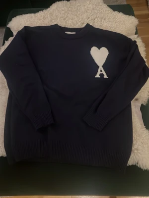 Mörkblå stickad tröja Ami Paris - Säljer en mörkblå stickad tröja från Ami Paris med deras ikoniska vita hjärta och A-logga på bröstet. Tröjan har rund halsringning, ribbade muddar och är långärmad. Perfekt för dig som gillar stilrena och trendiga plagg.