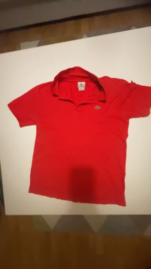 Röd piké från Lacoste, storlek S/M - Säljer en klassisk röd pikétröja från Lacoste med den ikoniska krokodilloggan broderad på bröstet. Tröjan har korta ärmar, krage och knappar vid halsen. Materialet är mjuk bomull som känns skönt mot huden. Perfekt för dig som gillar sportig och stilren look.