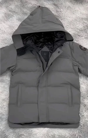 Grå dunjacka med huva från Canada Goose - Säljer en grå dunjacka från Canada Goose med stor huva och svarta detaljer på insidan. Jackan har två sidofickor, diskret logga på ärmen och dragkedja med knappslå framtill. Perfekt för kalla dagar och riktigt snygg stil.