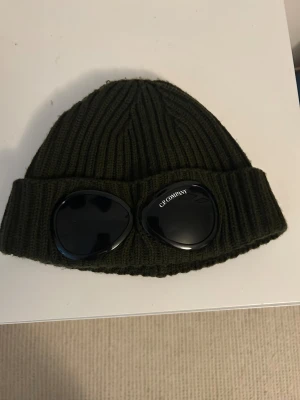 Mörkgrön C.P. Company goggle-mössa - Säljer en mörkgrön ribbstickad mössa från C.P. Company med ikoniska svarta goggle-linser framtill. Mössan är tillverkad i mjuk ull och har en bred uppvikt kant. Perfekt accessoar för dig som vill sticka ut och hålla dig varm.