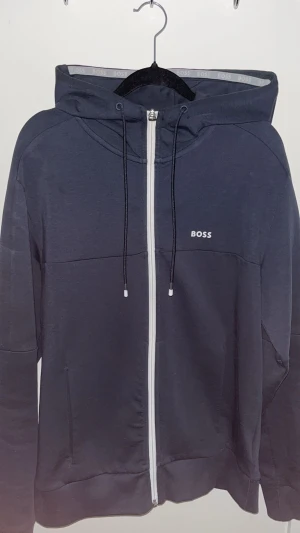 Mörkblå hoodie från BOSS med dragkedja - Snygg mörkblå kofta från BOSS med vit dragkedja och vita detaljer längs ärmarna. Tröjan har använd 2 gånger helt ny skick 