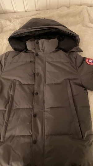Mörkgrå Canada Goose pufferjacka - Säljer en mörkgrå pufferjacka från Canada Goose med ikonisk röd och vit patch på ärmen. Jackan har huva, hög krage och stora fickor framtill. Stängs med både dragkedja och knappar. Perfekt för kalla dagar och riktigt snygg stil. Passar S och M