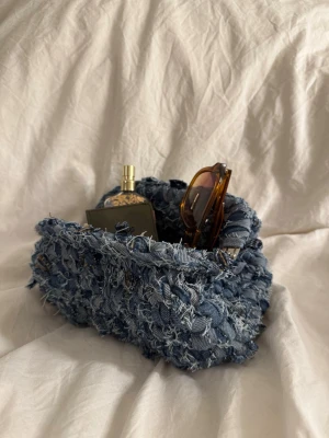 Blå handväska i denim med fransar - 016 - Blå. Unik handväska i blå denim med grov, flätad struktur och fransiga detaljer. Väskan har en mjuk, rundad form och är perfekt för dig som gillar en cool och kreativ stil. Materialet är återvunnen denim som ger en riktigt trendig vibe.