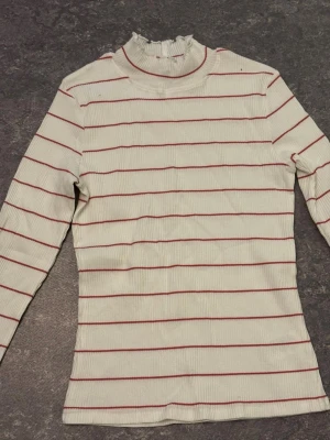 Vit och röd randig polotopp - Säljer en vit polotopp med röda horisontella ränder och ribbad struktur. Toppen har lång ärm och en hög krage med volangkant som ger en extra touch. Perfekt för dig som gillar klassiska mönster med en twist. Har dock några små fläckar som inte är så synliga