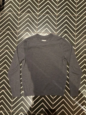 Mörkgrå superior merino från These Glory Days - En stilren mörkgrå stickad tröja med rund hals och långärmad passform. Tröjan är tillverkad i mjuk ull och har en klassisk, enkel design som funkar till många olika looks. Perfekt för dig som gillar minimalistisk stil och vill ha något som är både bekvämt och snyggt.