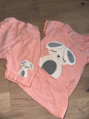 Rosa shorts med hundmotiv från Pompdelux Rosa Set med T-shirt och Shorts - Säljer ett par söta rosa shorts från Pompdelux med elastisk midja och broderat hundmotiv i vitt och grått på benet. Mjukt bomullsmaterial som är skönt för sommaren. Perfekt för små barn som gillar gulliga detaljer. Ett sött set bestående av en t-shirt och ett par shorts. Båda plaggen har en applikation av en sovande kanin.