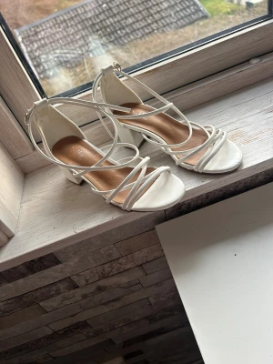 Vita klackar från Duffy - Säljer ett par vita klackar  från Duffy med blockklack och tunna remmar som korsar över foten. Skorna har öppen tå och spänne runt vristen. Perfekta för dig som gillar stilrena och trendiga skor med en feminin touch.