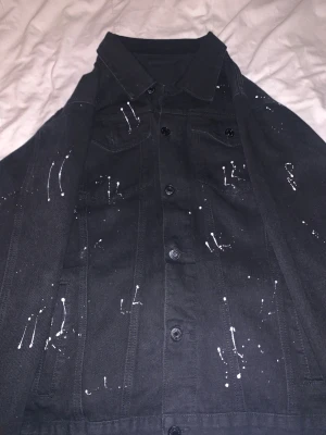 Svart jeansjacka med vit färgstänk - Svart jeansjacka med coola vita färgstänk över hela jackan. Klassisk krage, knäppning med svarta knappar och två bröstfickor. Jackan har en rak passform och är tillverkad i robust denim som ger en streetig vibe.