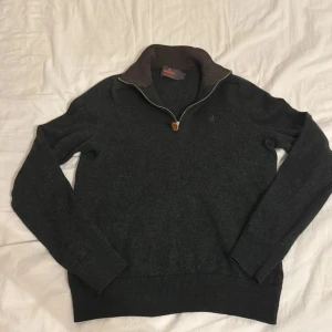 Mörkgrön merinoullströja från Morris - Snygg mörkgrå tröja från Morris i 100% merinoull med halv dragkedja och hög krage. Broderad logga på bröstet och ribbade muddar. Perfekt för lager-på-lager och chill höst- eller vinterstil