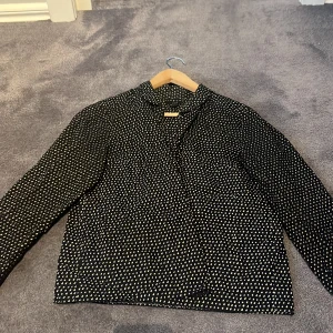 Svart prickig skjorta från COS - Svart skjorta från COS med beige prickigt mönster. Skjortan har lång ärm, klassisk krage och rak passform. Tillverkad i ett lätt och mjukt material som känns skönt mot huden. Perfekt för dig som gillar stilrena men ändå lekfulla plagg.