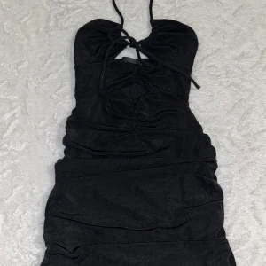 Svart kort klänning från Bershka - Säljer en svart kort klänning från Bershka med halterneck och snygg snörning framtill. Klänningen har rynkade detaljer och en liten cut-out. Materialet är stretchigt och formar kroppen.  Skriv om flera bilder önskas 🥰