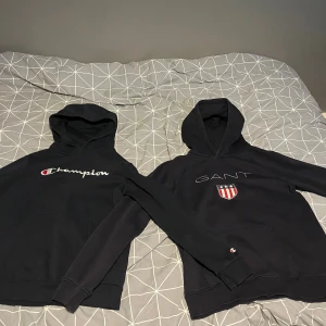 Svarta hoodies från Champion & GANT - Två svarta hoodies, en från Champion med broderad logga och en från GANT med broderad logga och sköld på bröstet. Båda har huva och magficka, perfekt för en chill och sportig stil.