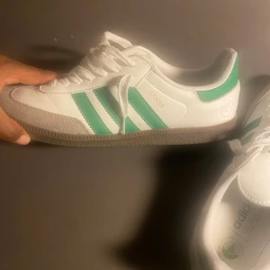 Adidas Samba vita och gröna sneakers - Säljer ett par klassiska Adidas Samba sneakers i vitt skinn med gröna ränder och detaljer. Skorna har snörning, grå mockadetaljer på tån och en brun gummisula. Perfekta för dig som gillar retrostil och vill ha något som sticker ut.