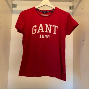 Röd GANT t-shirt med tryck - Röd t-shirt från GANT med vit text 'GANT 1949' på bröstet. Klassisk rund halsringning och korta ärmar. Skön passform och tillverkad i mjuk bomull, perfekt för en avslappnad stil.