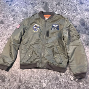 Grön bomberjacka Polo Ralph Lauren - Militärgrön bomberjacka från Polo Ralph Lauren med coola patches, orange foder och dragkedja framtill. Jackan har ribbade muddar, fickor med tryckknappar och en ärmficka med dragkedja. Storlek M (10-12 år).