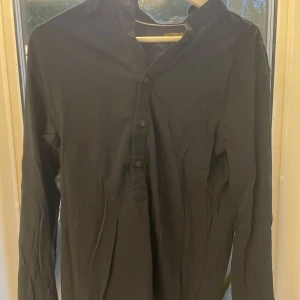 Svart henley från AllSaints Spitalfields - Svart henley-skjorta från AllSaints Spitalfields i storlek Large. Skjortan har lång ärm, klassisk krage och knappar framtill. Tillverkad i mjuk bomull som känns skön mot huden. Perfekt för dig som gillar stilrena och enkla plagg med snygg passform.