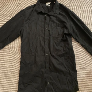 Svart skjorta från Jack & Jones - Svart långärmad skjorta från Jack & Jones Core i slim fit-modell. Skjortan har klassisk krage, bröstficka och knäppning framtill samt en snygg dragkedjedetalj på sidan. 