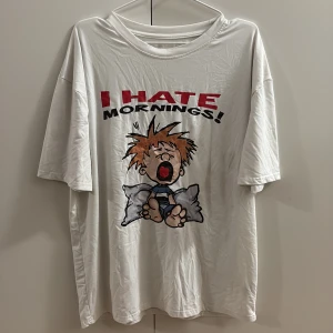 Vit t-shirt med tryck 'I Hate Mornings' - Rolig vit t-shirt med stort tryck på framsidan där det står 'I HATE MORNINGS!' och en tecknad figur som ser trött ut. T-shirten har korta ärmar och en loose passform, perfekt för chill dagar. Materialet känns mjukt och bekvämt mot huden.