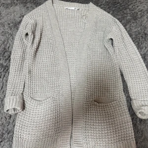 Beige stickad kofta med fickor - Mysig beige stickad kofta från H&M med grovstickat mönster och två praktiska fickor framtill. Koftan har lång ärm och ribbade muddar, perfekt att slänga på sig när det är lite kyligare ute.