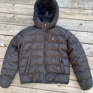 Svart pufferjacka från Gant med huva - Svart jacka från GANT med avtagbar huva. Fickor med tryckknapp i sidan för säker förvaring och resår i nederkant och ärmslut för en bra passform. Printad logga på vänster bröst. En varm jacka som står emot vind bra, perfekt för kalla dagar.