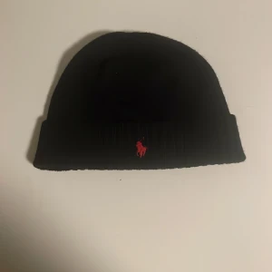 Svart ullmössa från Polo Ralph Lauren - Svart mössa i 100% ull från Polo Ralph Lauren. Klassisk ribbstickad design med uppvikt kant och röd broderad Polo-logga framtill. Perfekt accessoar för kalla dagar och snygg till streetwear eller casual outfits.