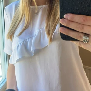Vit blus med volangdetalj - Säljer en vit blus med korta ärmar och snygg volangdetalj framtill. Blusen har en rak passform och är tillverkad i ett lätt, mjukt material som ger en fräsch och clean look. Perfekt för dig som gillar stilrena plagg med en twist.