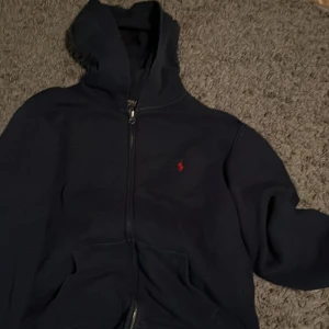Mörkblå hoodie från Polo Ralph Lauren - Mörkblå hoodie från Polo Ralph Lauren med dragkedja framtill och klassisk röd logga broderad på bröstet. Tröjan har huva, kängurufickor och långa ärmar. Perfekt för chill dagar eller när du vill ha en clean streetstyle.
