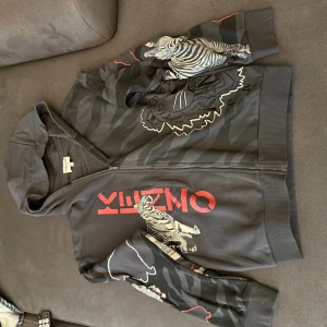 Kenzo hoodie med djurprint och huva - Säljer en svart Kenzo hoodie med dragkedja och huva. Tröjan har grått zebramönster och stora tryck av elefanter, tigrar och giraffer i beige och vitt. Röd Kenzo-logga framtill. Perfekt statement-plagg för dig som gillar unika prints.