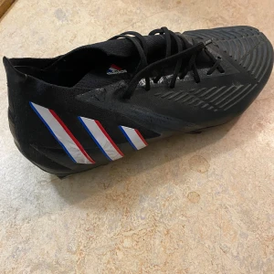 Adidas Predator svarta fotbollsskor - Säljer ett par svarta Adidas Predator fotbollsskor med klassiska tre ränder i vitt, rött och blått på sidan. Skorna har snörning, mönstrad ovansida för bättre bollkontroll och en strumpliknande passform. Perfekta för dig som vill dominera på planen.