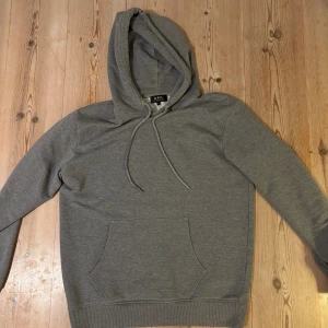 Grå hoodie från A.P.C. med huva - Säljer en stilren grå hoodie från A.P.C. med klassisk känguruficka och dragsko i huvan. Tröjan har lång ärm och är tillverkad i mjuk bomull, perfekt för en chill och avslappnad look.
