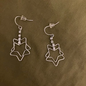 Silvriga örhängen med stjärnor - Säljer ett par coola silvriga örhängen med dubbla stjärnformer och gnistrande stenar på hängen. Perfekt för dig som vill sticka ut med en unik och glittrig detalj till din outfit. Ej riktigt silver och köpta i Japan💖🇯🇵