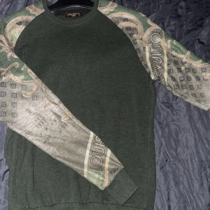 Grön Louis Vuitton tröja med mönstrade ärmar - Säljer en grön tröja från Louis Vuitton med unika långärmade ärmar i grått och grönt, dekorerade med klassiskt LV-mönster och detaljerade tryck. Tröjan har rund halsringning och en stilren passform som sticker ut.