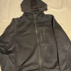 Svart Sail Racing Hoodie - Svart hoodie från Sail Racing med dragkedja framtill och huva. Snygg Sail Racing-logga på bröstet och stor broderad text på ryggen. Praktisk bröstficka med dragkedja. Perfekt för dig som gillar sportig och clean stil.