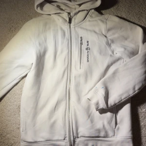 Vit hoodie från Sail Racing med dragkedja - Vit hoodie från Sail Racing med hel dragkedja, broderad logga på bröstet och praktisk bröstficka med dragkedja. Mjuk insida och huva med broderad detalj. Perfekt för chill dagar eller när du vill ha en clean look.