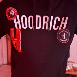 Svart hoodie från Hoodrich med tryck - Svart hoodie från Hoodrich med stora vita och röda tryck och broderade patches framtill. Tröjan har huva med snörning och känguruficka. Perfekt för dig som gillar streetwear och vill sticka ut med en fet logga på bröstet.
