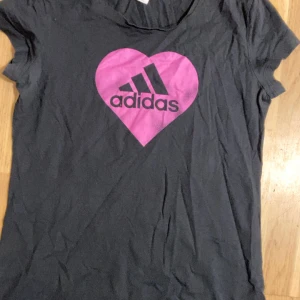 Svart Adidas t-shirt med rosa hjärta - Svart t-shirt från Adidas med korta ärmar och ett stort rosa hjärta med Adidas-logga på bröstet. Skön passform och tillverkad i mjuk bomull. Perfekt för dig som gillar sportiga och snygga plagg med en färgklick.