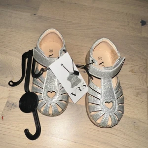 Glittriga silversandaler med hjärtdetalj - Säljer ett par silvriga sandaler från VRS med glittrigt material och en söt hjärtformad detalj framtill. Sandalerna har öppna tår, kardborreband och platt sula. Perfekta för sommarens alla äventyr och riktigt snygga till både shorts och klänning.