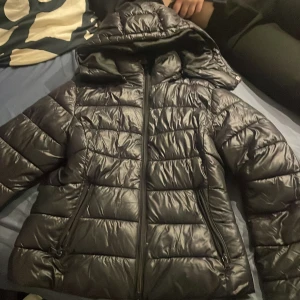 Svart pufferjacka med huva - Säljer en svart pufferjacka med huva och dragkedja framtill. Jackan har ett quiltat mönster och två sidofickor med dragkedja. Perfekt för kalla dagar och har en snygg, lite glansig finish. Passar dig som gillar en stilren och praktisk look.