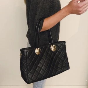 Vera Pelle väska - En svart vera Pelle väska med snygga guldfärgade detaljer vid handtagen. Perfekt storlek för att få plats med allt du behöver och ger en stilren vibe till din outfit. 