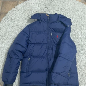 Blå dunjacka Polo Ralph Lauren S - Säljer en marinblå dunjacka från Polo Ralph Lauren med röd broderad logga på bröstet. Jackan har hög krage, dragkedja framtill och flera fickor med dragkedja. Perfekt för kalla dagar och riktigt snygg streetstil.