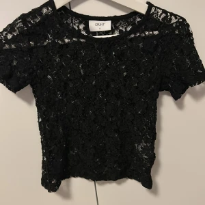 Svart spetsblus från Grunt - Säljer en svart blus från Grunt i storlek S. Blusen är helt i spets med korta ärmar och rund hals. Den har ett transparent och detaljerat mönster som ger en cool och edgy vibe. Perfekt för dig som gillar att sticka ut med din stil.