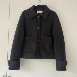 Svart ulljacka från Nelly - Stilren svart jacka från Nelly i ullblandning med klassisk krage, stora fickor framtill och knäppning med knappar. Jackan har långa ärmar med snygga detaljer vid ärmsluten.