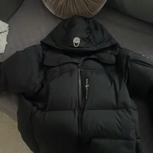 Svart pufferjacka Sail Antarctica - Svart pufferjacka från Sail Antarctica Expedition med stor huva och coola logodetaljer. Jackan har dragkedja framtill och är rejält vadderad för att hålla värmen. Perfekt för kalla dagar och har en clean, modern look.